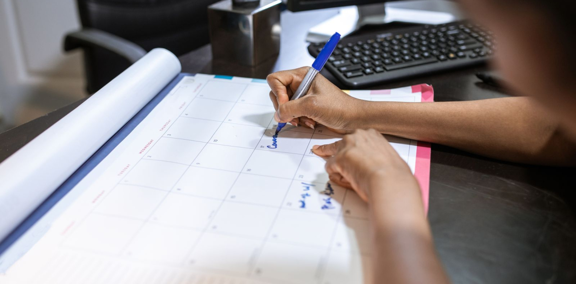 Prise de rendez-vous : une main inscrit une date sur un calendrier de bureau, devant un ordinateur