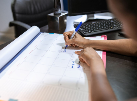 Prise de rendez-vous : une main inscrit une date sur un calendrier de bureau, devant un ordinateur